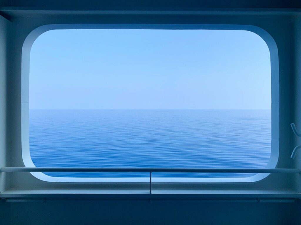 A calm blue ocean viewed through a rectangular ship window with a railing